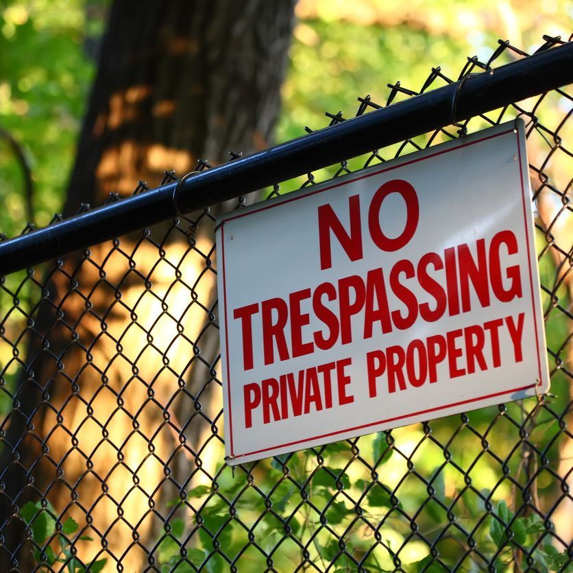 A close-up image of a 'No trespassing - private property' sign hanging on a fence with green trees on the background